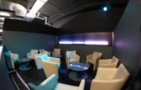 Warehouse, © Warehouse Production GmbH Lounge-Bereich mit beigen Sesseln und runden Tischen in einem modernen Raum.