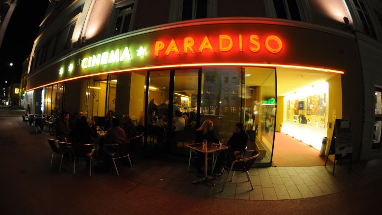 Cinema Paradiso, © Andrea Reischer Exterior view of a movie theater called Cinema Paradiso at night with illuminated entrance and people at tables.