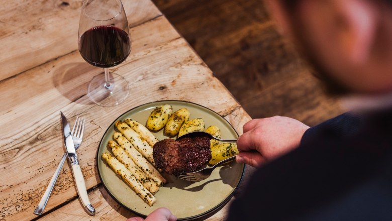 Fillet of beef roasted on a hot stone, © Niederösterreich Werbung/David Schreiber A plate with fillet of beef, asparagus and potatoes, next to a glass of red wine on a wooden table.