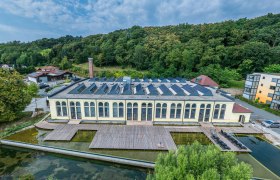 Spitzenfabrik, © Sebastian Wegerbauer Luftaufnahme einer alten Fabrik mit Solarpanelen auf dem Dach, umgeben von Bäumen und einem Teich.