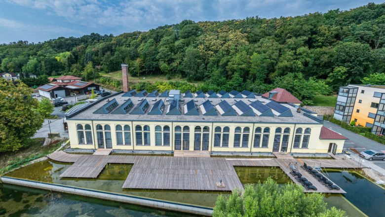 Spitzenfabrik, © Sebastian Wegerbauer Luftaufnahme einer alten Fabrik mit Solarpanelen auf dem Dach, umgeben von Bäumen und einem Teich.