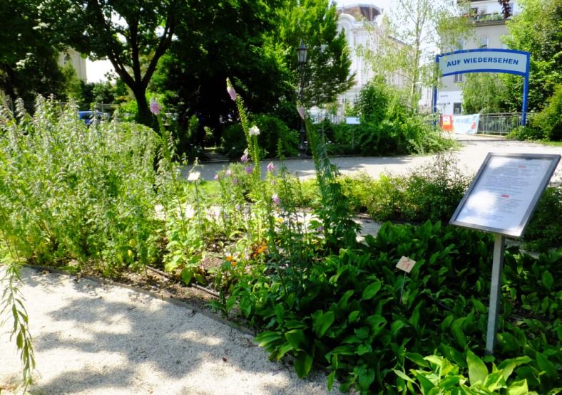 Ein gepflegter Garten mit Pflanzen und einem Schild, im Hintergrund ein Tor mit der Aufschrift 'Auf Wiedersehen'.