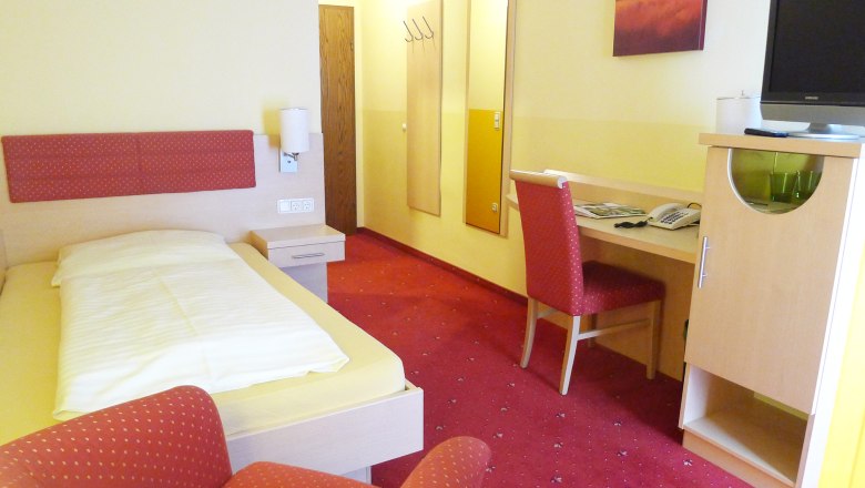 Ein Hotelzimmer mit Einzelbett, rotem Teppich, Schreibtisch und Stuhl. An der Wand hängt ein Bild, daneben ein Fernseher auf einem Schrank.