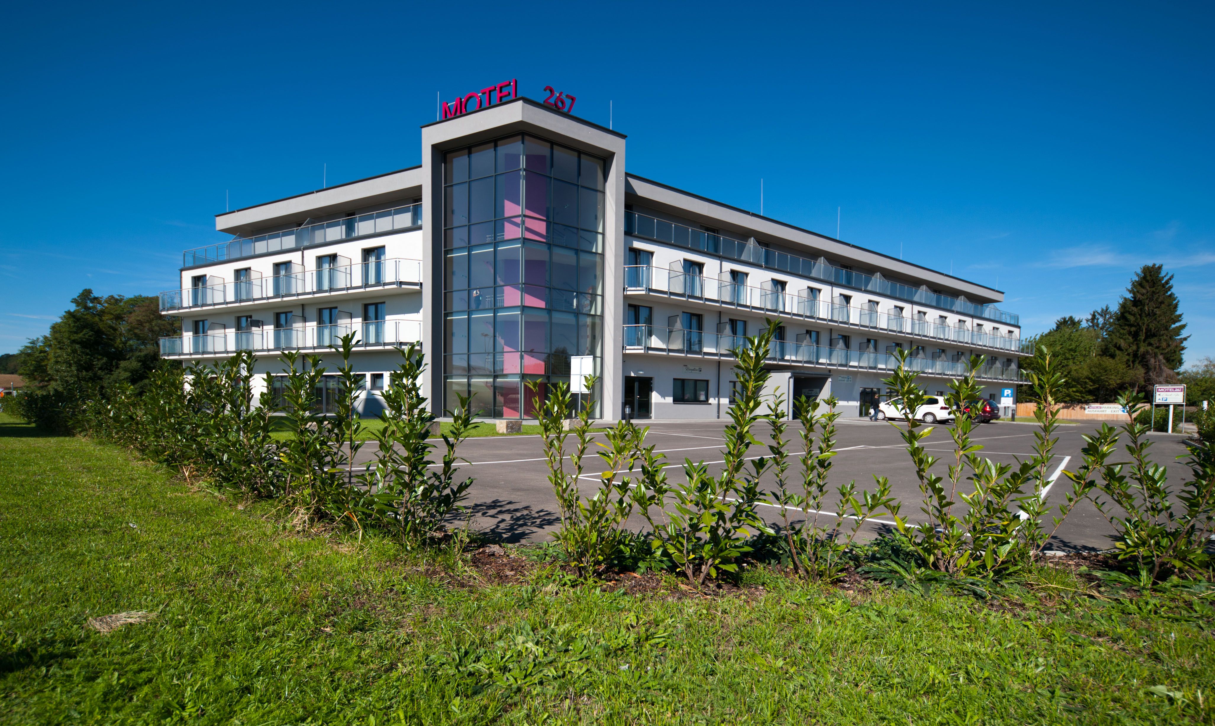 Modernes dreistöckiges Motel mit Glasfassade und Parkplatz, umgeben von grüner Landschaft.