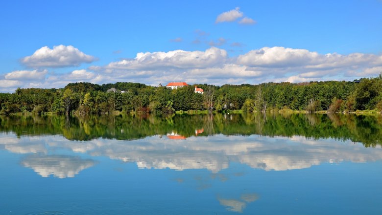 A lake with a reflection of trees and clouds, in the background a building with a red roof.