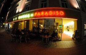 Außenansicht eines Kinos namens Cinema Paradiso bei Nacht mit beleuchtetem Eingang und Menschen an Tischen.
