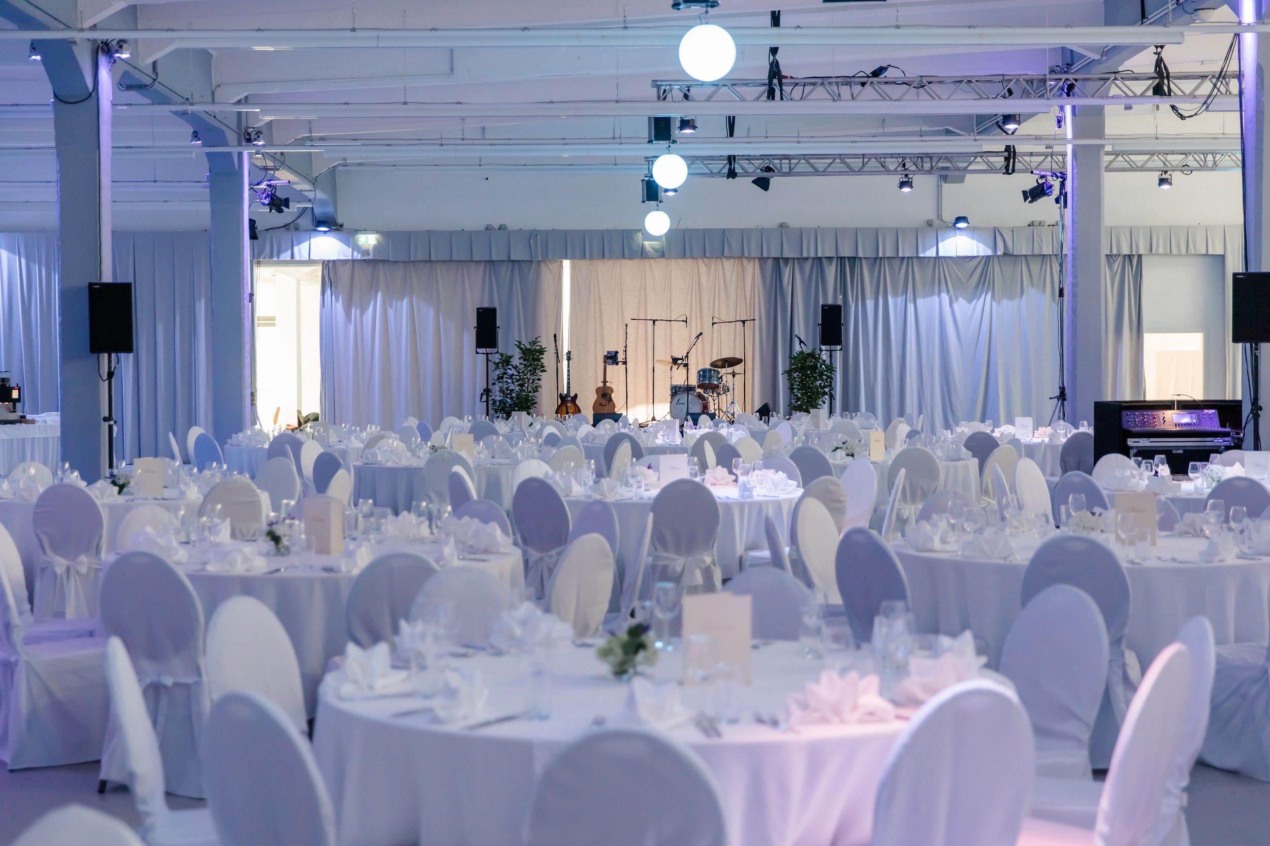 Eleganter Veranstaltungsraum mit runden, weiß gedeckten Tischen und Bühne mit Musikinstrumenten.