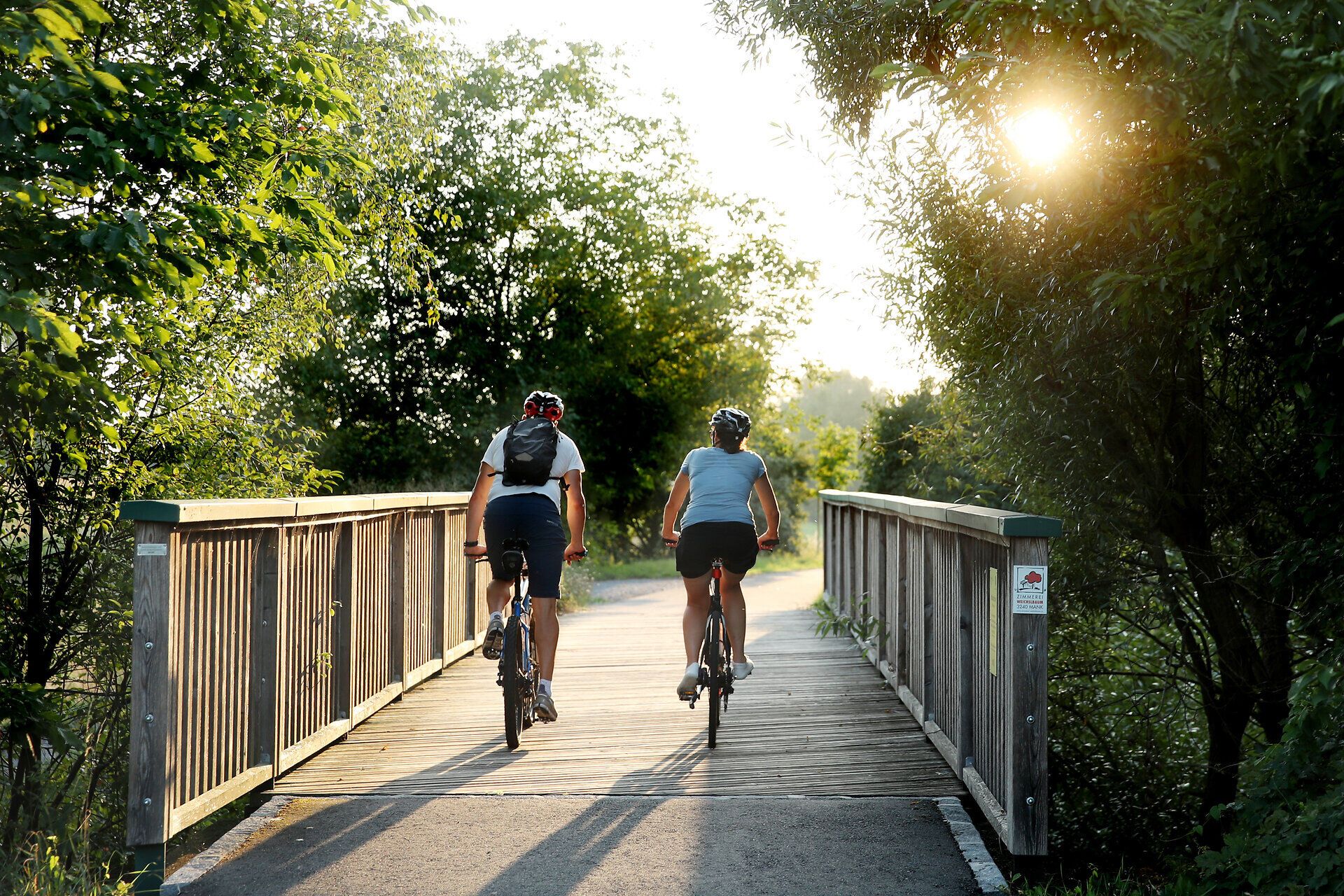 Zwei Radfahrer genießen die sanfte Brise und die warmen Sonnenstrahlen, während sie über eine malerische Holzbrücke fahren. Umgeben von üppigem Grün und dem sanften Licht des Sommers, lädt die idyllische Landschaft zu unvergesslichen Erlebnissen im Mostviertel ein.