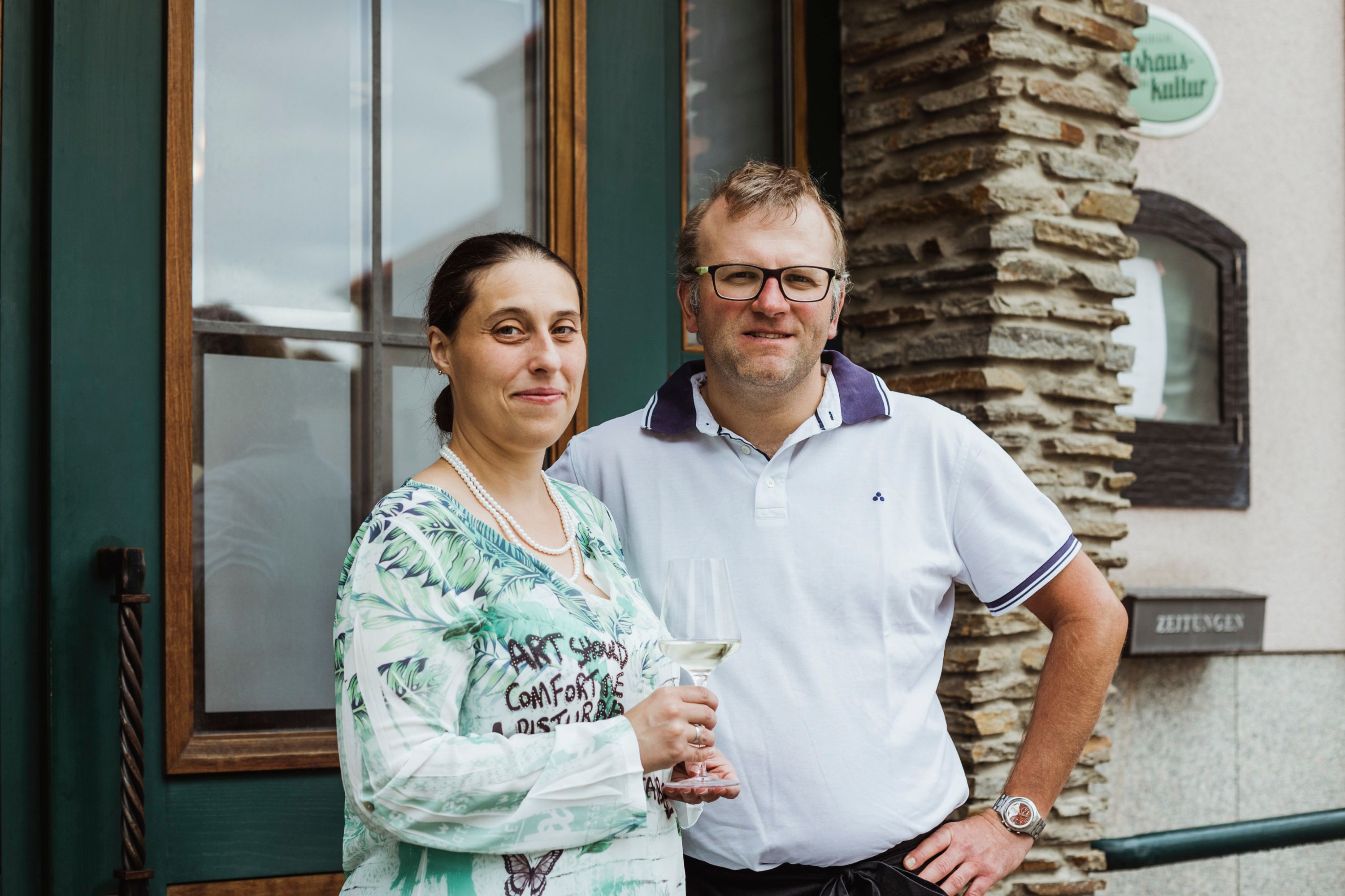 A man and a woman stand smiling in front of a building. The woman is holding a wine glass.