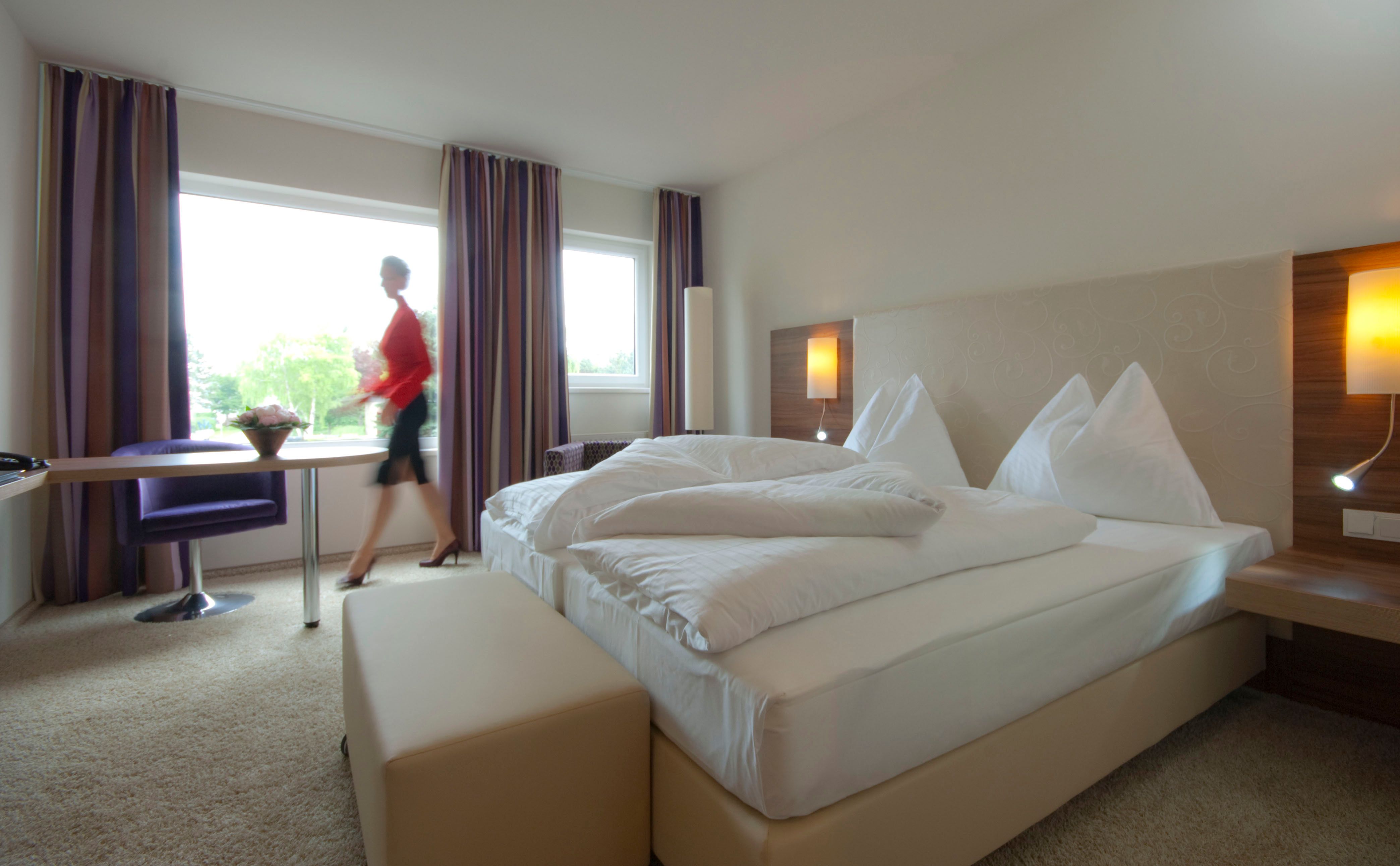 Modern eingerichtetes Hotelzimmer mit Doppelbett, großem Fenster und einer Person, die vorbeigeht.