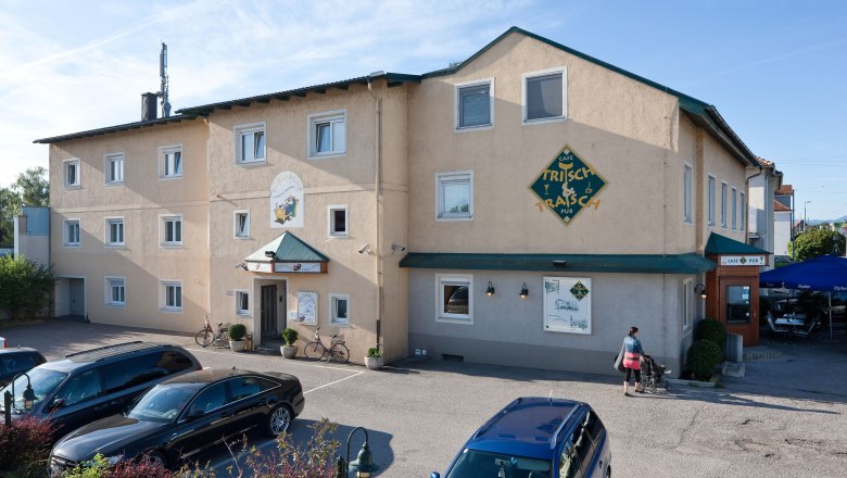 Au&szlig;enansicht eines Geb&auml;udes mit der Aufschrift 'Pension Schlafgut' und 'Caf&eacute; Trisch & Tratsch'. Autos parken davor, eine Person mit Kinderwagen geht vorbei.