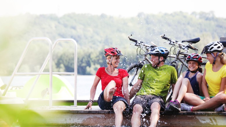 Gruppe von Menschen mit Fahrradhelmen sitzt lachend auf einem Steg am Wasser.