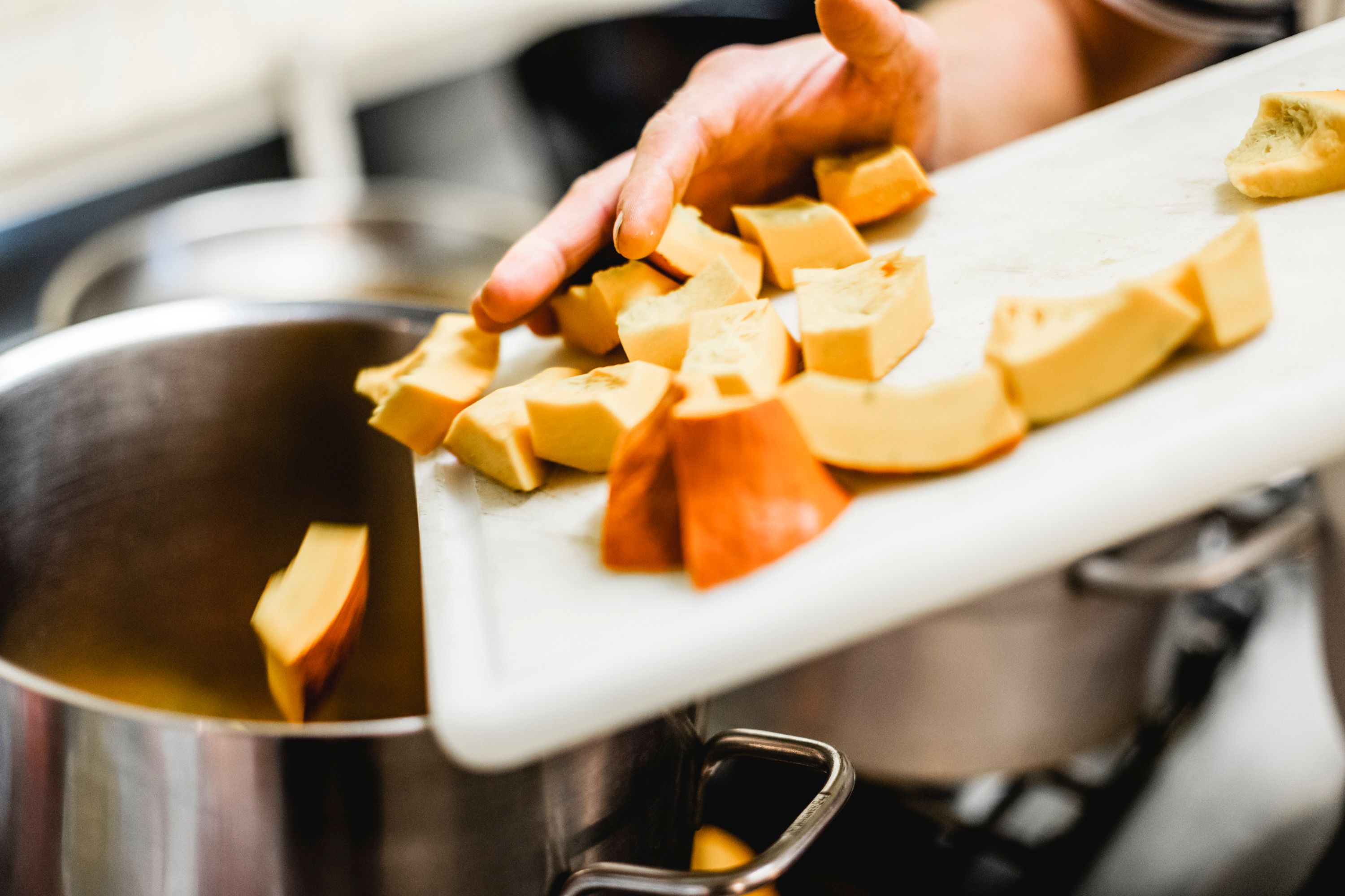 A hand pushes sliced pumpkin pieces from a cutting board into a pot.