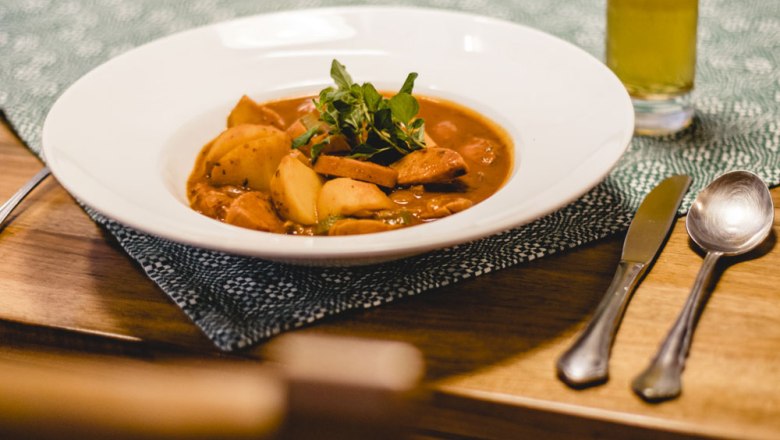 Ein Teller mit Kartoffelgulasch auf einem Tisch, daneben Besteck und ein Glas Getr&auml;nk.