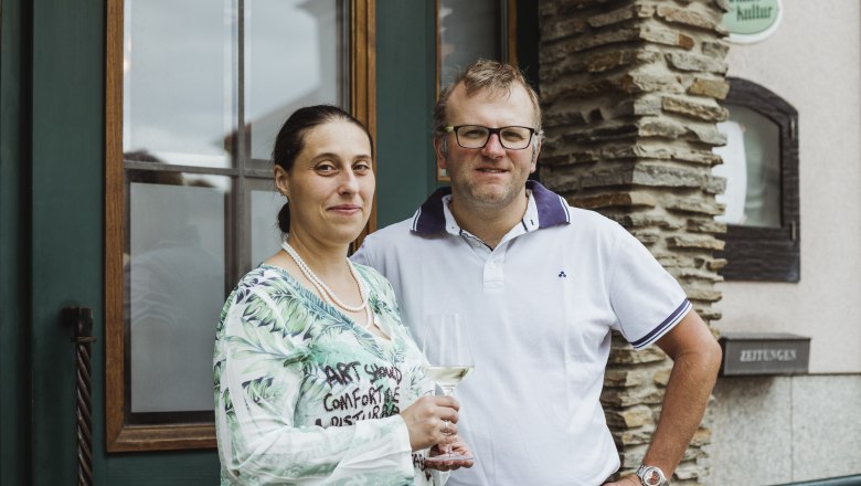 Ein Mann und eine Frau stehen l&auml;chelnd vor einem Geb&auml;ude. Die Frau h&auml;lt ein Weinglas in der Hand.