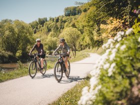 Two cyclists ride along the Traisental cycle route, which leads through wooded areas.