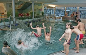Kinder spielen und planschen in einem Hallenbad mit Wasserspielen.