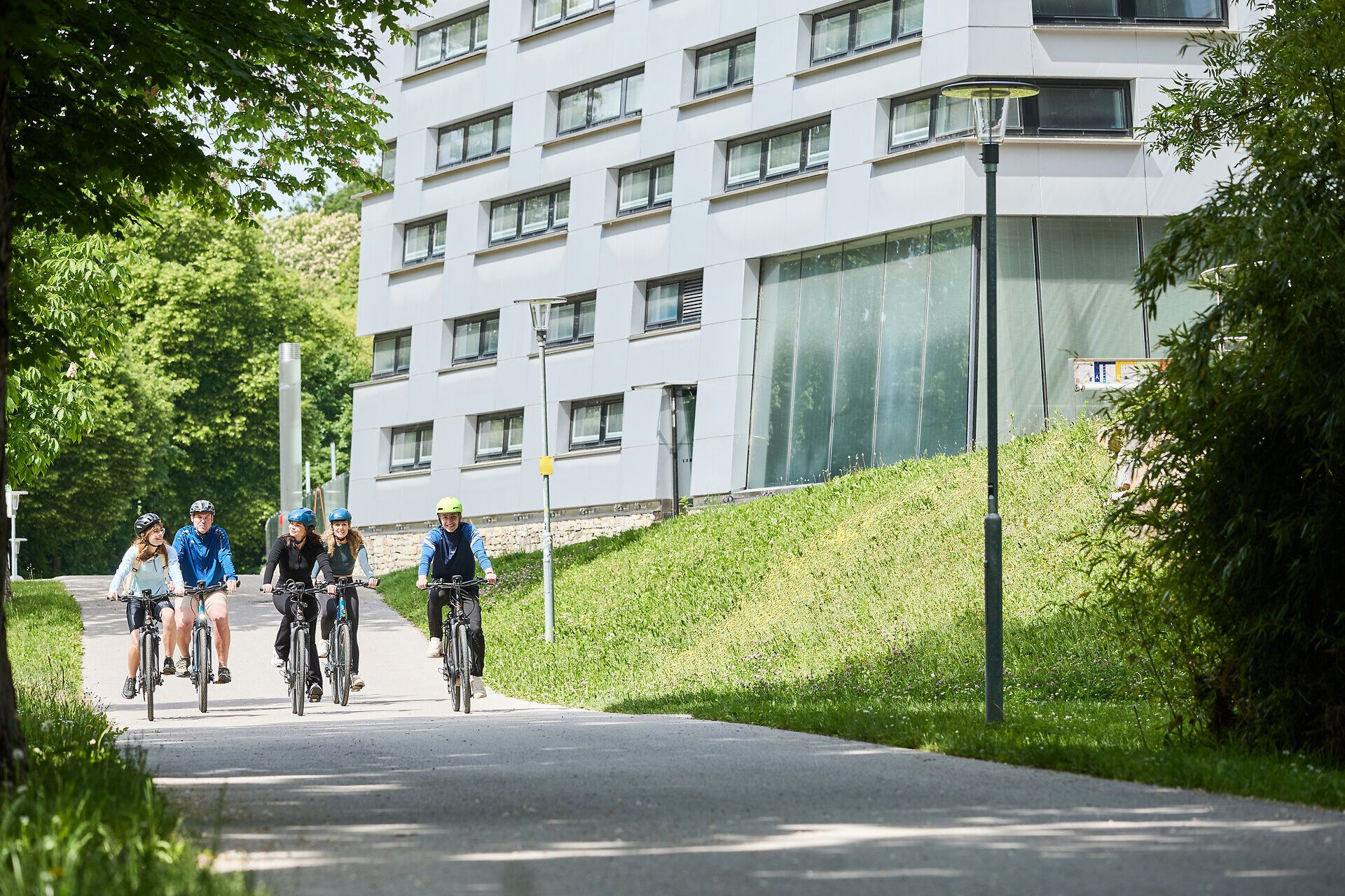 Radfahrende in Parkanlage mit Bäumen und Blick auf zeitgenössische Architektur.