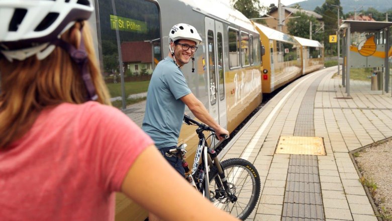 Zwei Personen mit Fahrradhelmen schieben ihre R&auml;der am Bahnsteig, neben ihnen steht die Himmelstreppe auf den Gleisen.