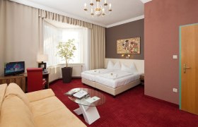 Hotelzimmer mit Doppelbett, rotem Teppich, Sofa, Fernseher und gro&szlig;em Fenster.