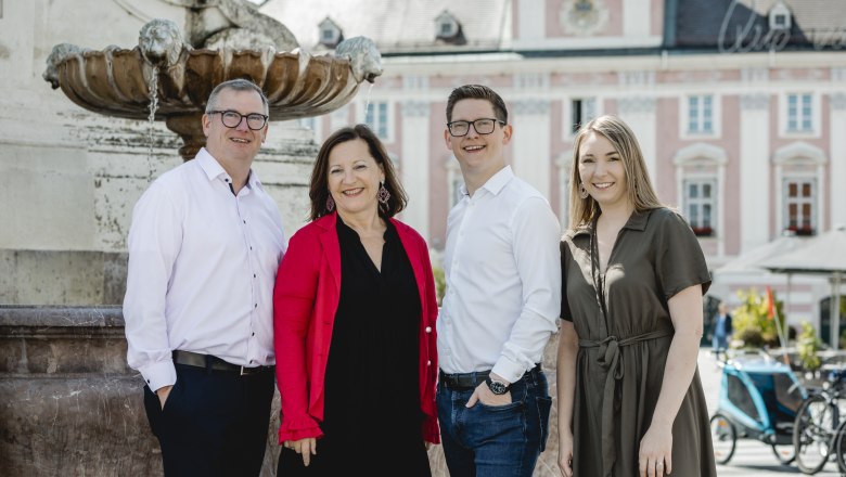 Four people stand smiling in front of a fountain in an urban setting.