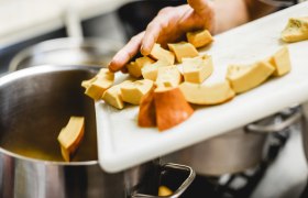 Eine Hand schiebt geschnittene K&uuml;rbisst&uuml;cke von einem Schneidebrett in einen Topf.