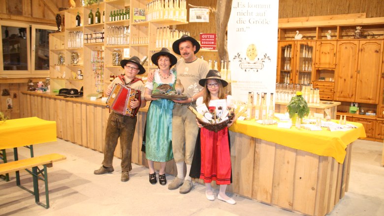 Eine Familie in traditioneller Kleidung steht in einem Laden mit Produkten und einem Banner mit der Aufschrift 'Die Wachtelei'.