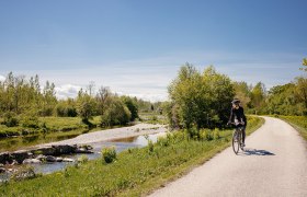 Ein malerischer Radweg schl&auml;ngelt sich entlang eines sanft pl&auml;tschernden Baches, umgeben von &uuml;ppigem Gr&uuml;n und bl&uuml;henden Wiesen. Die klare Luft und die strahlende Sonne laden dazu ein, die Sch&ouml;nheit der Natur zu genie&szlig;en und die Seele baumeln zu lassen.