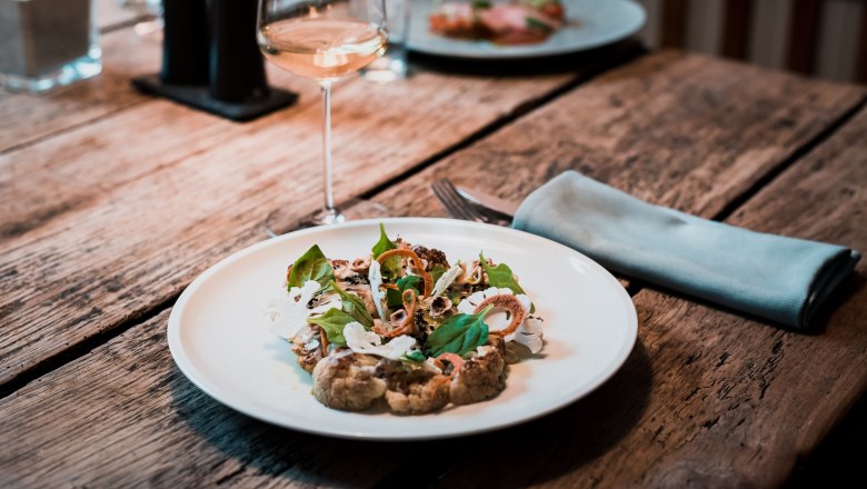 Ein Teller mit geschmortem Karfiol, Miso Spinat und eingelegtem Sanddorn auf einem Holztisch, daneben ein Glas Weißwein.