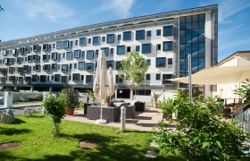 Modernes Geb&auml;ude mit vielen Fenstern und Gartenbereich mit Sitzgelegenheiten.