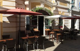 Außenbereich eines Restaurants mit Tischen, Stühlen und Sonnenschirmen in einer sonnigen Gasse.
