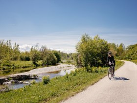 Ein malerischer Radweg schl&auml;ngelt sich entlang eines sanft pl&auml;tschernden Baches, umgeben von &uuml;ppigem Gr&uuml;n und bl&uuml;henden Wiesen. Die klare Luft und die strahlende Sonne laden dazu ein, die Sch&ouml;nheit der Natur zu genie&szlig;en und die Seele baumeln zu lassen.