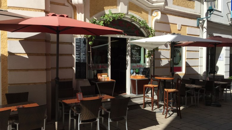 Außenbereich eines Restaurants mit Tischen, Stühlen und Sonnenschirmen in einer sonnigen Gasse.