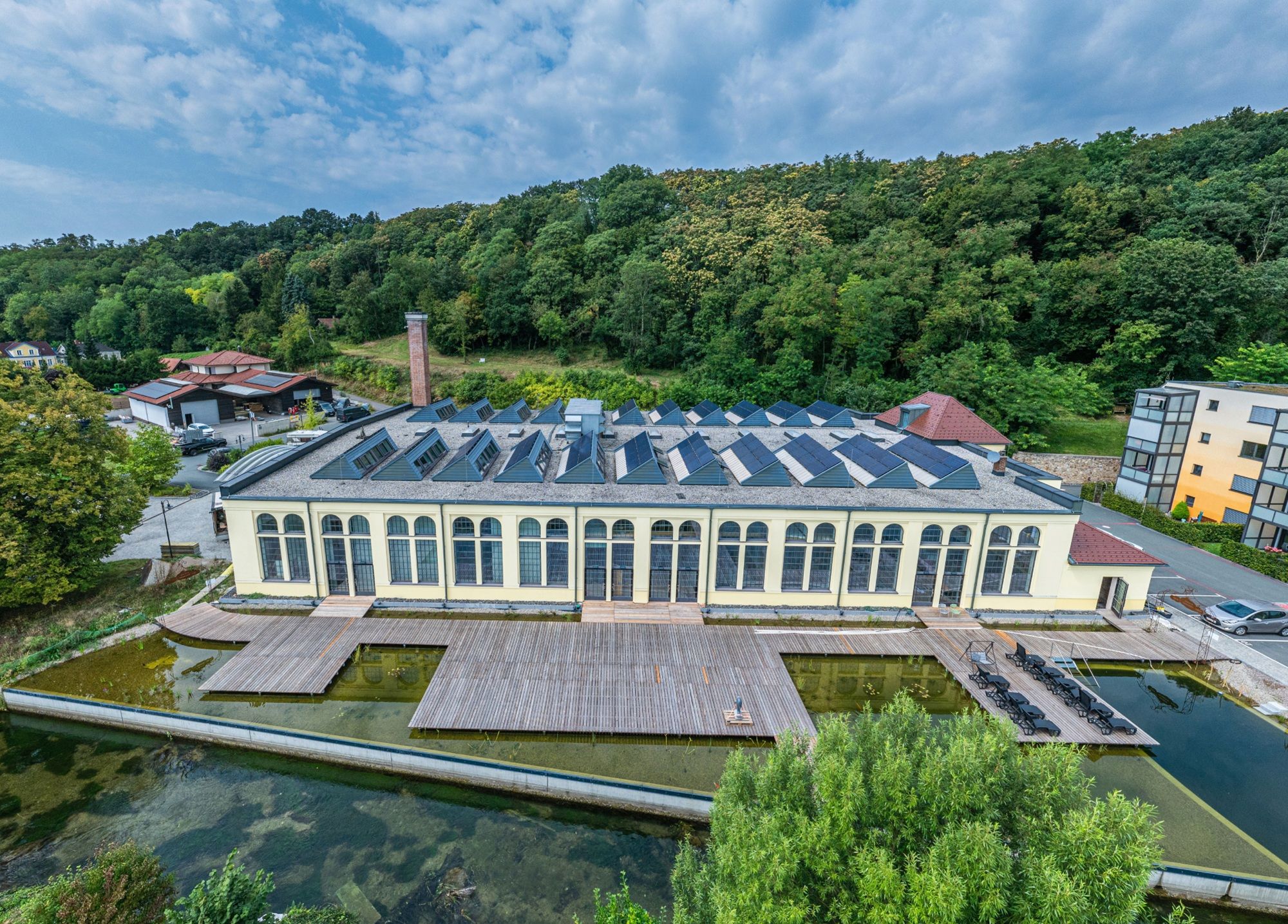 Luftaufnahme einer alten Fabrik mit Solarpanelen auf dem Dach, umgeben von Bäumen und einem Teich.