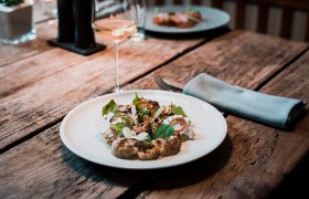 Ein Teller mit geschmortem Karfiol, Miso Spinat und eingelegtem Sanddorn auf einem Holztisch, daneben ein Glas Weißwein.