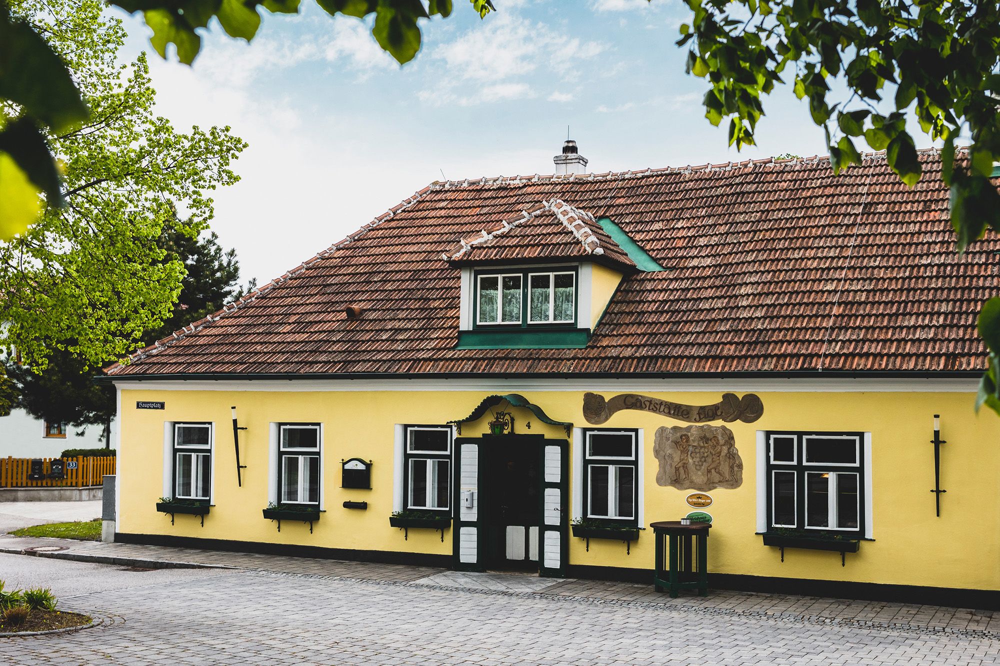 Gelbes Gebäude mit roten Dachziegeln und grünen Fensterläden, umgeben von Bäumen.